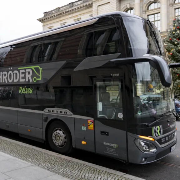 (3)	Grauer Doppeldecker-Reisebus von „Schröder Reisen“ steht am Straßenrand vor einem historischen Gebäude; daneben ist ein geschmückter Weihnachtsbaum zu sehen.