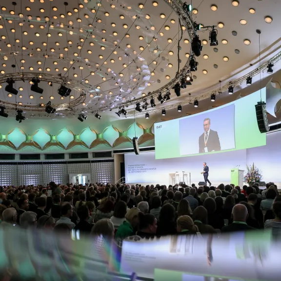 Großer Konferenzsaal mit Bühne, Redner und aufmerksamem Publikum, moderne Beleuchtung