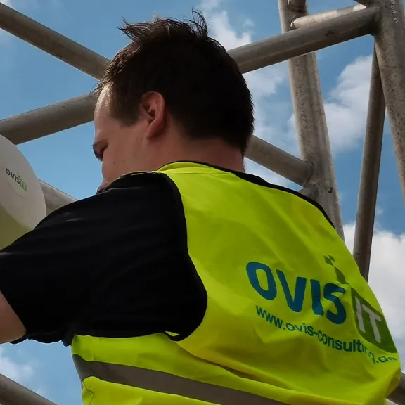 Techniker mit gelber Weste montiert Gerät an Metallgerüst bei blauem Himmel.