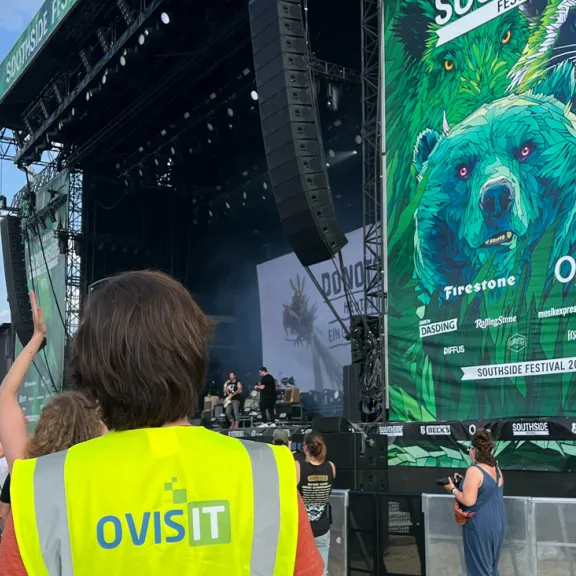 Mensch mit gelber Weste bei Open-Air-Konzert, große Bühne im Hintergrund.