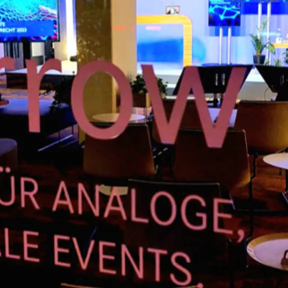 Blick durch Glas mit Schrift auf modernen Veranstaltungsraum mit Bildschirmen und Sitzplätzen.