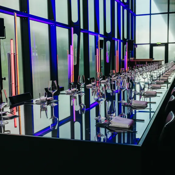 Langer, moderner Esstisch mit Gläsern und Besteck, beleuchtet von bunten Lichtern.