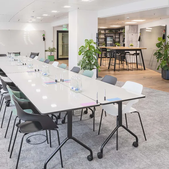 Heller Konferenzraum mit langem Tisch, vielen Stühlen und Pflanzen, moderne Einrichtung.