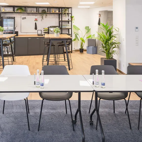 Moderner Konferenzraum mit weißen Tischen, Stühlen und Pflanzen, im Hintergrund eine offene Küche.