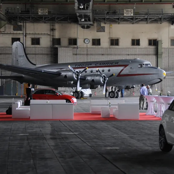 Silbernes Flugzeug in Hangar, rote Teppichfläche mit Autos und Stehtischen davor