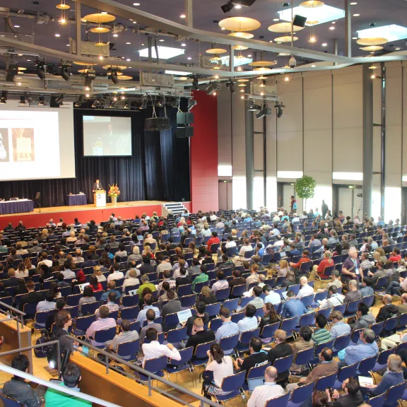 Großer Konferenzsaal mit vielen sitzenden Teilnehmern und einer Präsentation auf der Bühne.