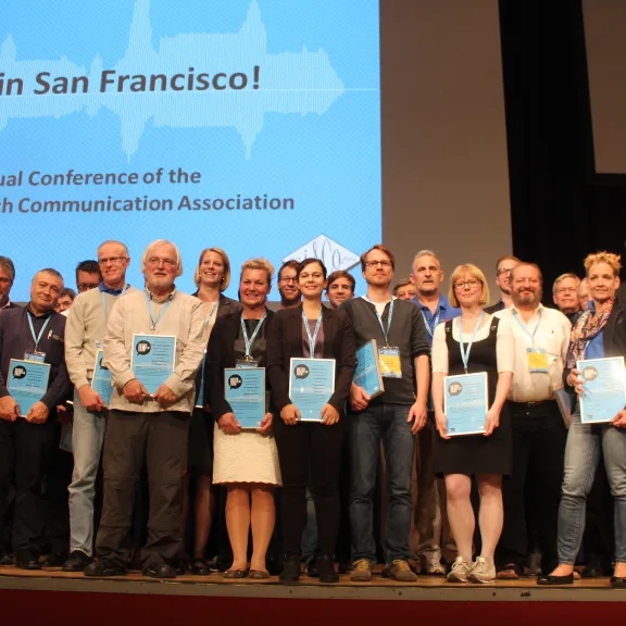 Gruppenfoto von Konferenzteilnehmern mit Urkunden auf einer Bühne.