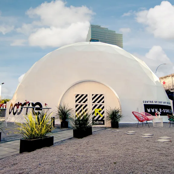 Weißes Kuppelzelt mit Pflanzen und bunten Stühlen auf einem Platz, blauem Himmel im Hintergrund.