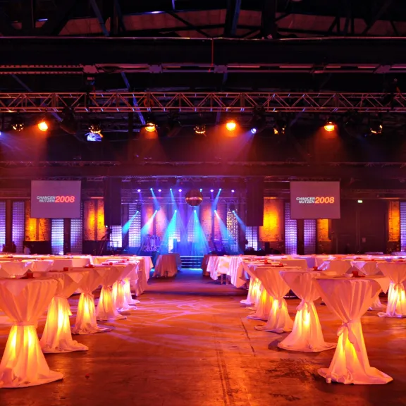 Festlich beleuchteter Saal mit Stehtischen und Bühne im Hintergrund.