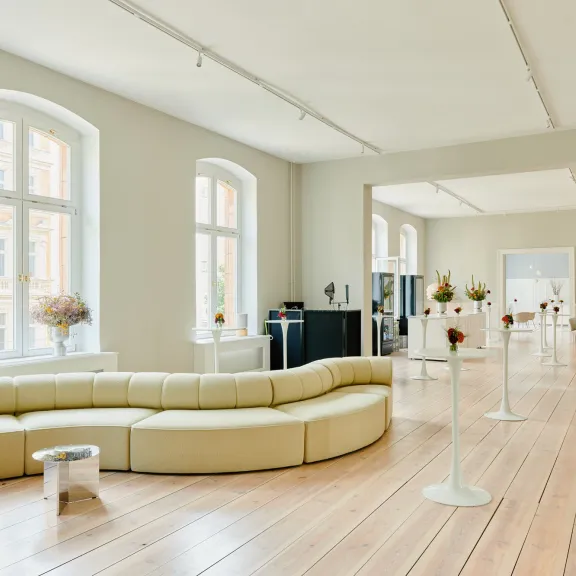 Heller & geräumiger Altbau-Saal mit Holzdielen-Boden, eingerichtet mit einem großes Design Sofa und Stehtischen mit kleiner Blumendekoration  