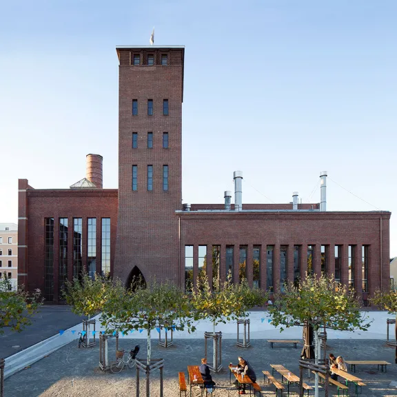 Backstein-Gebäude mit hohem Turm, davor Platz mit Bäumen und Sitzbänken.