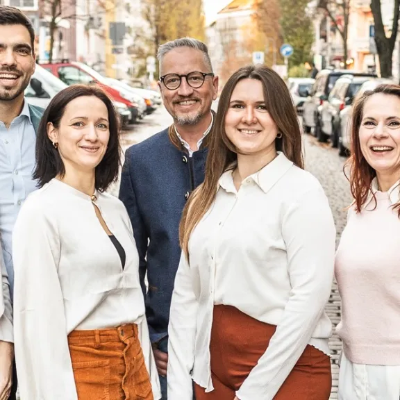 Neun lächelnde Menschen stehen auf einer sonnigen Straße in einer Stadt.