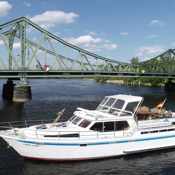 Weißes Motorboot auf Fluss vor grüner Stahlbrücke bei sonnigem Wetter.