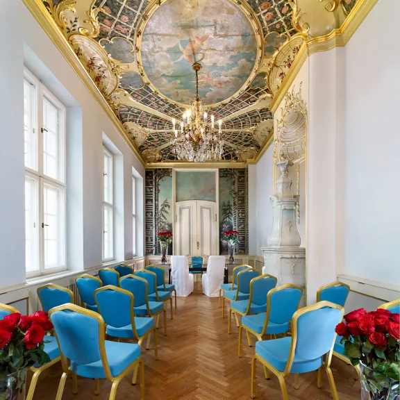 Prunkvoller Hochzeitssaal mit blauen Stühlen, Kronleuchter und bemalter Decke.