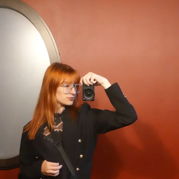Frau mit rotem Haar macht ein Spiegelselfie vor roter Wand.
