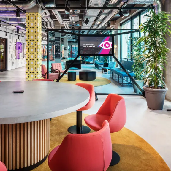 Modernes Büro mit runden Tischen, roten Stühlen, Pflanzen und großer Fensterwand.