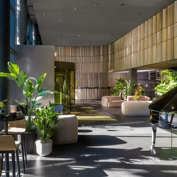 Heller Hotellobby mit großen Fenstern, Pflanzen, Sofas und einem schwarzen Flügel