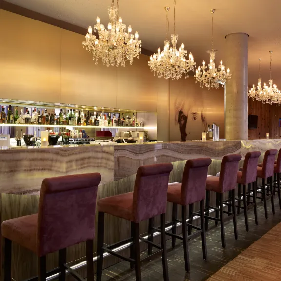 Moderne Bar mit lila Barhockern, beleuchteten Kronleuchtern und gut sortierten Regalen.