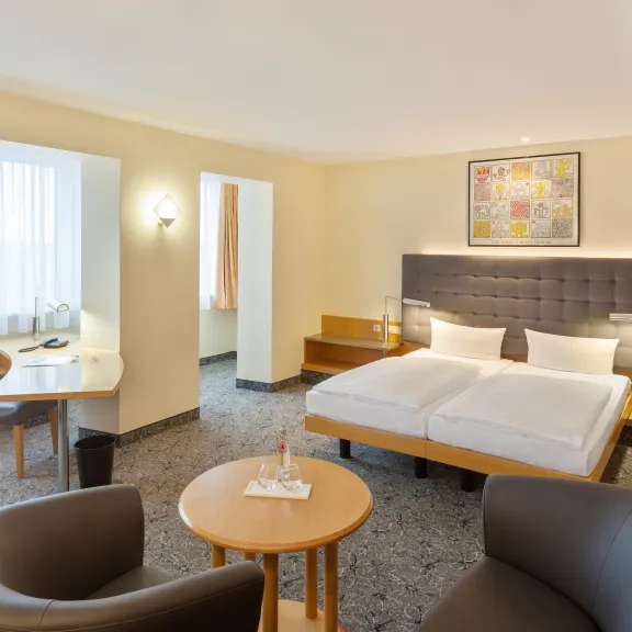 Helles Hotelzimmer mit Doppelbett, Sitzbereich und Schreibtisch, modern eingerichtet.