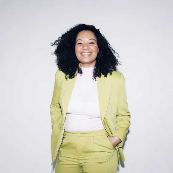 ein Foto von der Gründerin und Geschäftsführerin von The Impact Company, Yolanda Rother, in einem grünen Anzug, lächelnd, sie steht vor einer weißen Wand