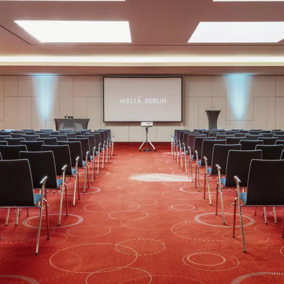 Konferenzraum mit roten Teppich, vielen Stuhlreihen und einer großen Leinwand vorne.