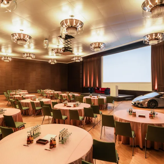 Konferenzraum mit runden Tischen, großer Leinwand und einem silbernen Auto auf der Bühne.