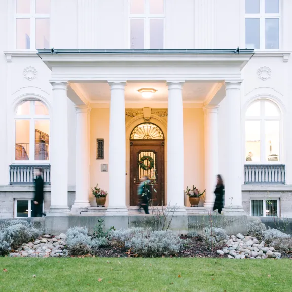 Weiße Villa mit Säulen, beleuchtetem Eingang und drei Personen im Vorgarten.