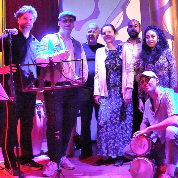 Gruppe von sieben Menschen auf einer Bühne, bunt beleuchtet, zwei halten Trommeln.