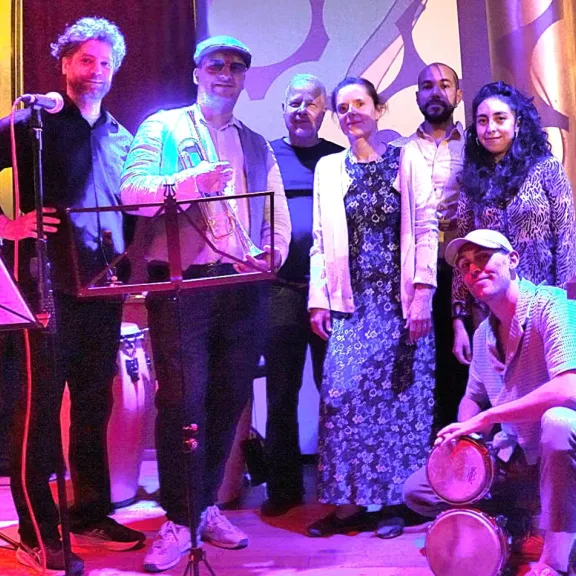 Gruppe von sieben Menschen auf einer Bühne, bunt beleuchtet, zwei halten Trommeln.