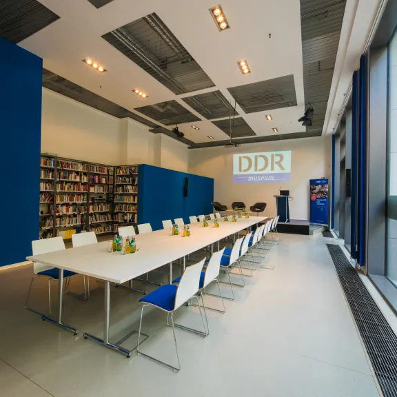 Moderner Konferenzraum mit langer Tafel, blau-weißen Stühlen und Bücherregalen.