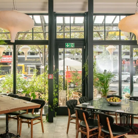Modernes Café mit großen Fenstern, Holztischen, Pflanzen und hellen Lampen.