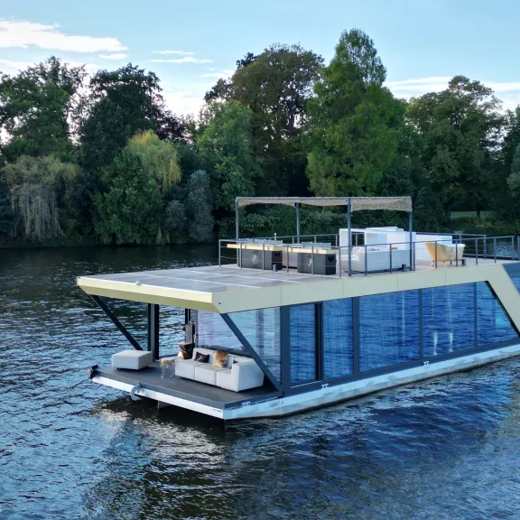 Modernes Hausboot mit Glasfront auf einem Fluss, umgeben von Bäumen.