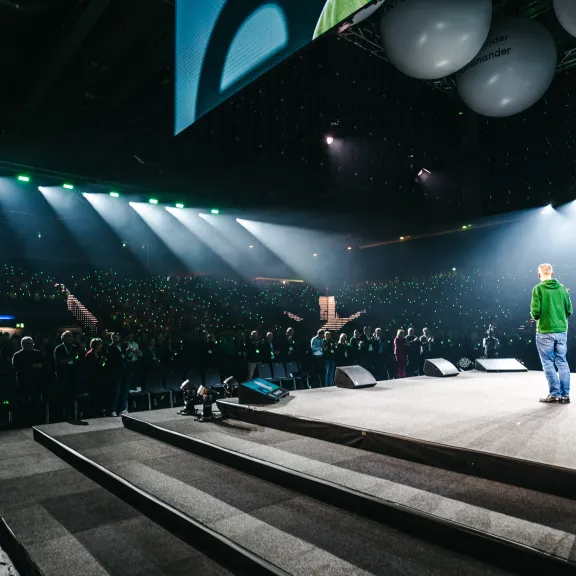 Person auf Bühne spricht vor großem, beleuchtetem Publikum in Arena.