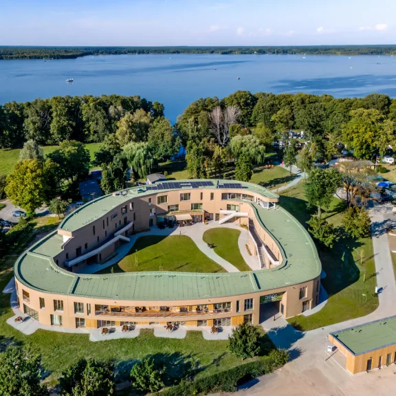 Modernes, ovales Gebäude mit begrüntem Dach am See, von Bäumen umgeben.