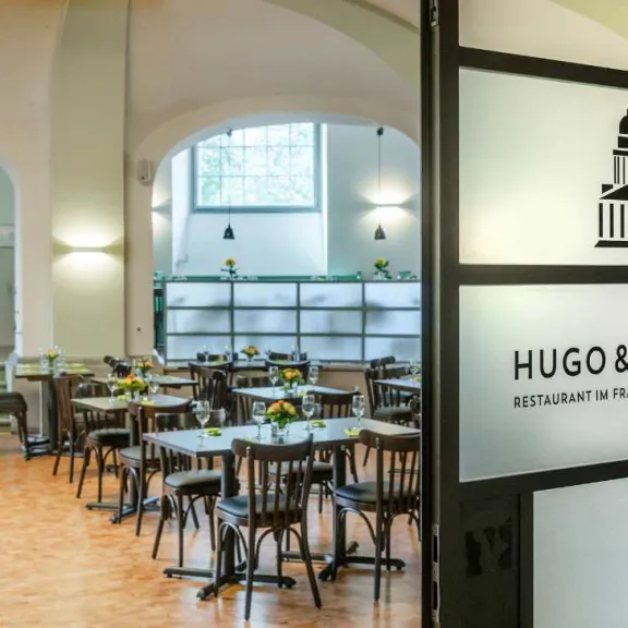 Modernes, helles Café mit Holztischen und Blumen, Eingang mit Logo „Hugo & Notte“.