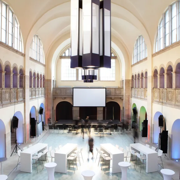 Großer, heller Veranstaltungssaal mit hohen Decken, Stehtischen und Leinwand.