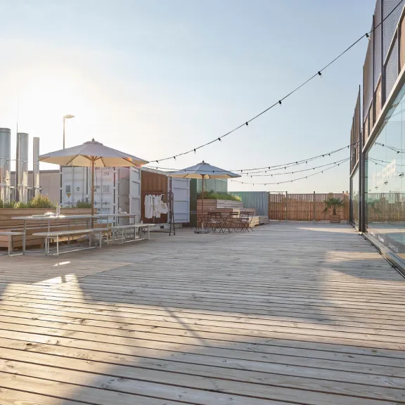 Sonnige Dachterrasse mit Holzboden, Tischen, Sonnenschirmen und Lichterketten.