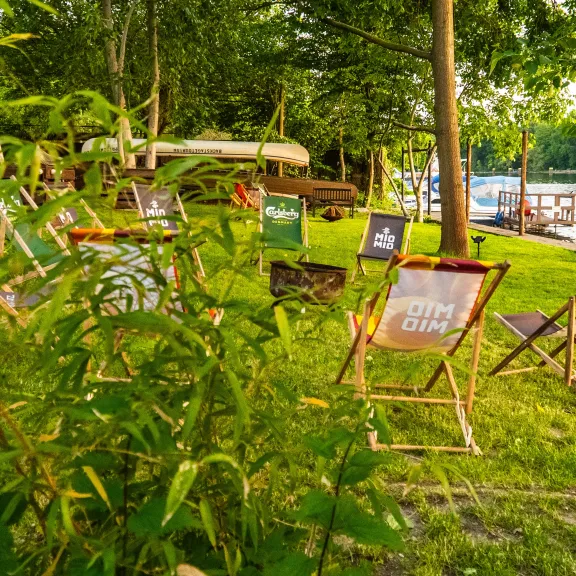 Mehrere Liegestühle stehen auf einer grünen Wiese am Seeufer im Sonnenschein.