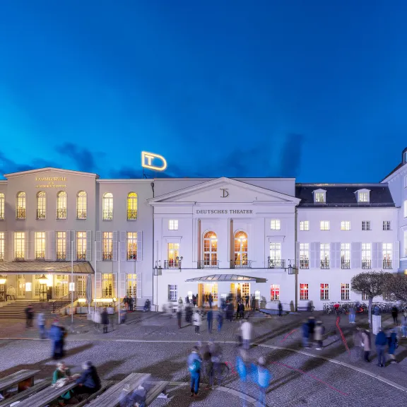 Beleuchtetes Theatergebäude abends mit Menschen auf dem Platz davor.
