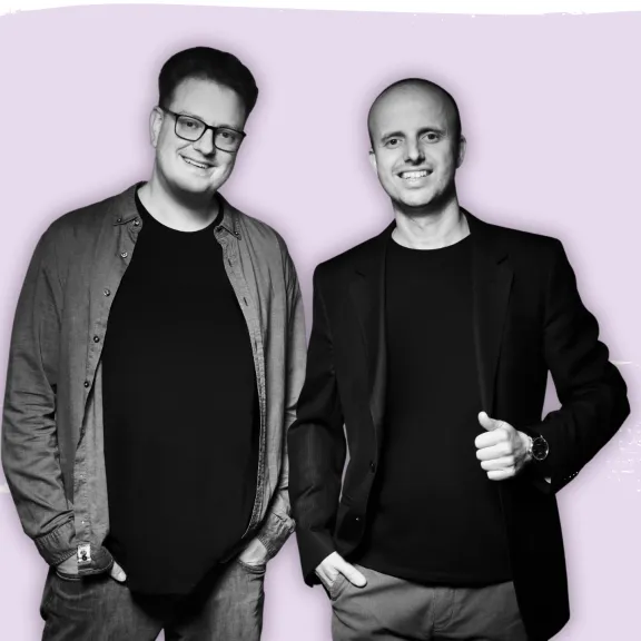 Zwei lächelnde Männer in dunkler Kleidung vor lila Hintergrund, links ein Event DJs Logo.