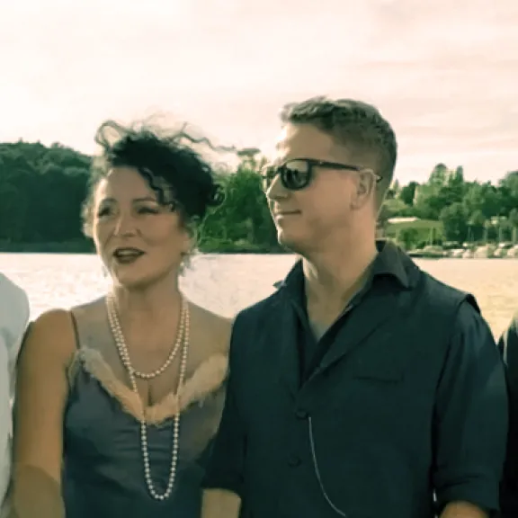 Vier Erwachsene stehen lächelnd am Wasser, sommerliche Kleidung, Bäume im Hintergrund.