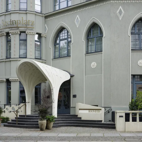 Gebäude mit rundem Vordach, großen Fenstern und Blumenkübeln am Eingang.
