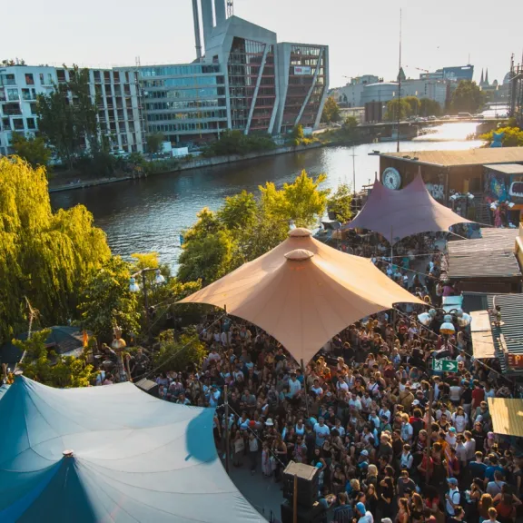 Belebtes Open-Air-Gelände vom Holzmarkt mit Zeltdächern und Menschenmenge am Spreeufer in der Abendsonne, im Hintergrund Stadtgebäude.