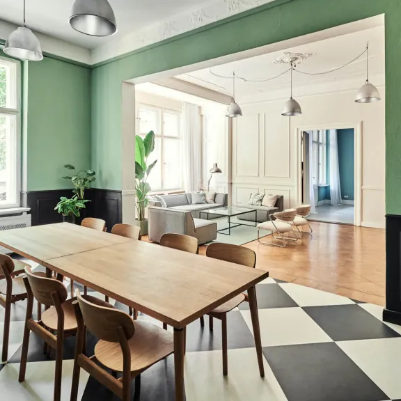 Großer heller Raum mit langem Holztisch, Stühlen und Schachbrettboden, angrenzendes Wohnzimmer.