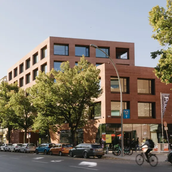 Modernes Backsteingebäude an einer Straße mit Bäumen, Autos und einem Radfahrer.