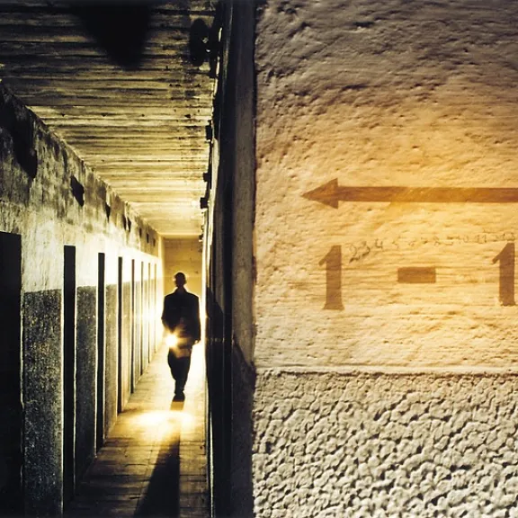 Dunkler Gang mit vielen Türen, eine silhouettierte Person mit Lichtquelle im Hintergrund.