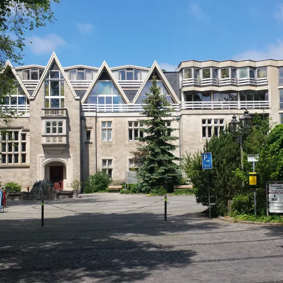 Historisches Gebäude mit spitzem Dach, Bäumen und blauem Himmel im Hintergrund.