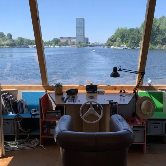 Steuerstand eines Bootes mit Ausblick auf einen Fluss und einen fernen Turm bei Sonnenschein.