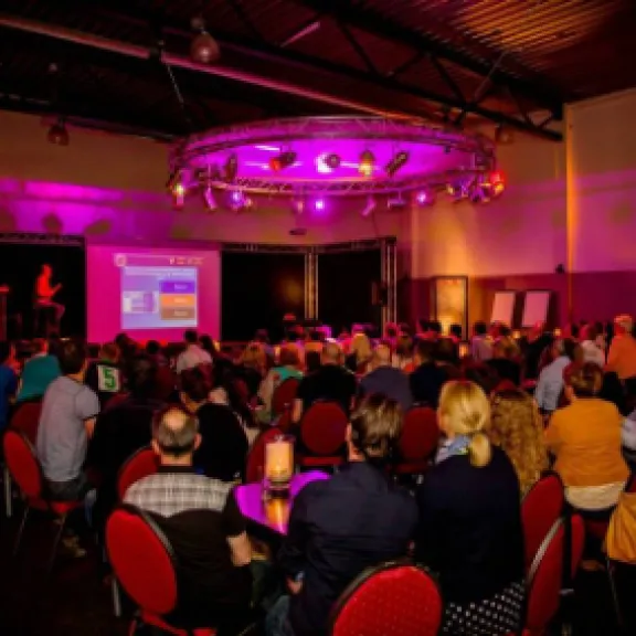 Viele Menschen sitzen im abgedunkelten Raum und sehen eine Präsentation auf Leinwand.
