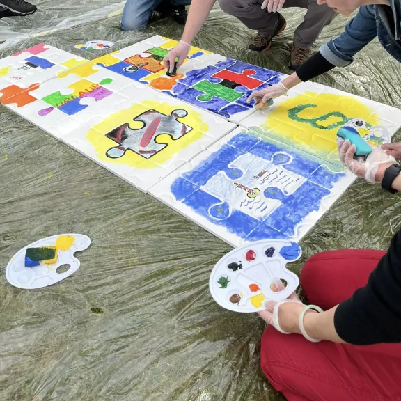 Mehrere Personen bemalen gemeinsam ein großes Puzzle-Bild mit bunten Farben.